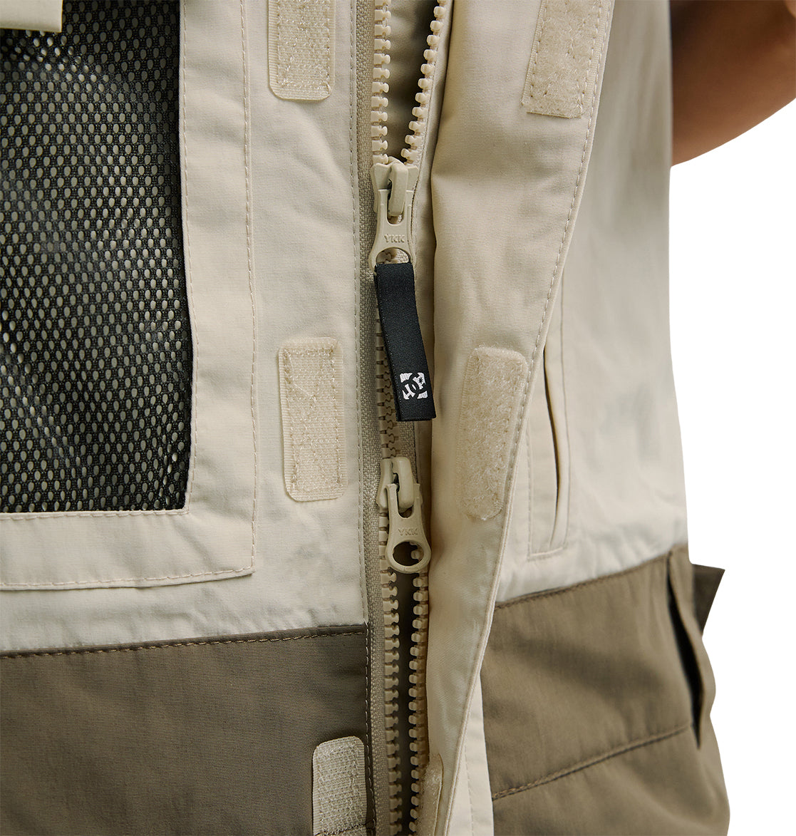 Close-up of the Mountain Lab Bib 20K by DC Shoes in beige and olive, featuring a partially unzipped zipper, mesh pocket, fabric tabs, and a black pull tab with small white branding—engineered for breathable protection and superior snow performance.