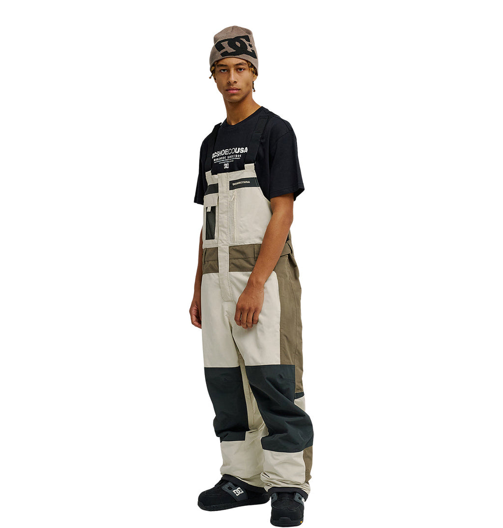 Wearing a graphic T-shirt, the individual is styled in DC Shoes’ Mountain Lab Bib 20K color-blocked overalls in beige, tan, and gray—designed for breathable protection—paired with black shoes and a brown beanie, standing slightly turned against a plain white backdrop.