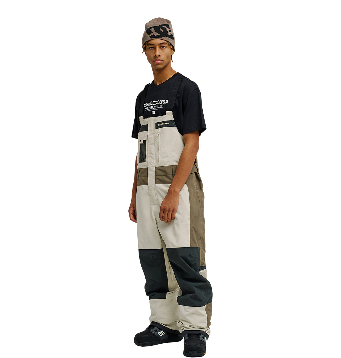 Wearing a graphic T-shirt, the individual is styled in DC Shoes’ Mountain Lab Bib 20K color-blocked overalls in beige, tan, and gray—designed for breathable protection—paired with black shoes and a brown beanie, standing slightly turned against a plain white backdrop.