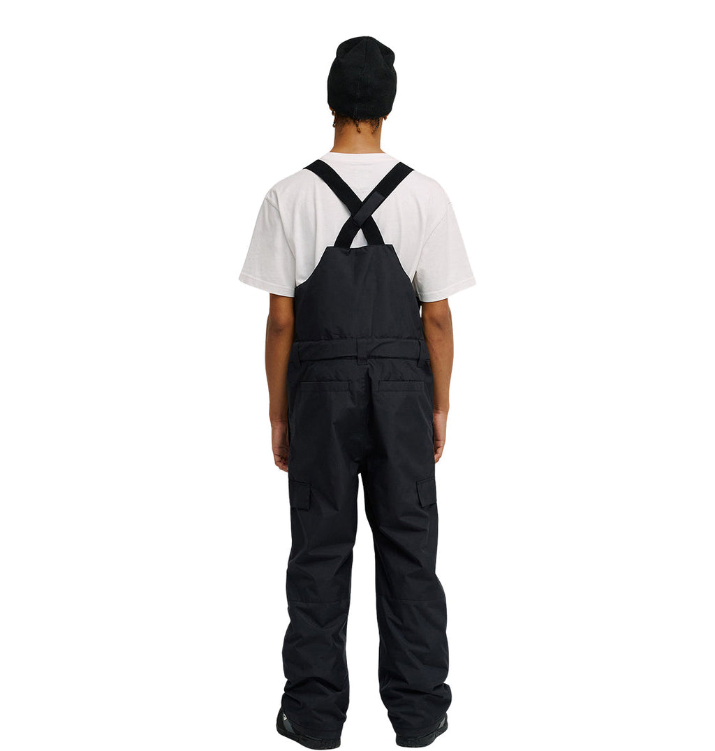 A person stands facing away against a plain white background, wearing a white t-shirt, black shoes, a black beanie, and the DC Shoes Docile Bib 10K—black insulated bib overalls featuring crisscross straps on the back.