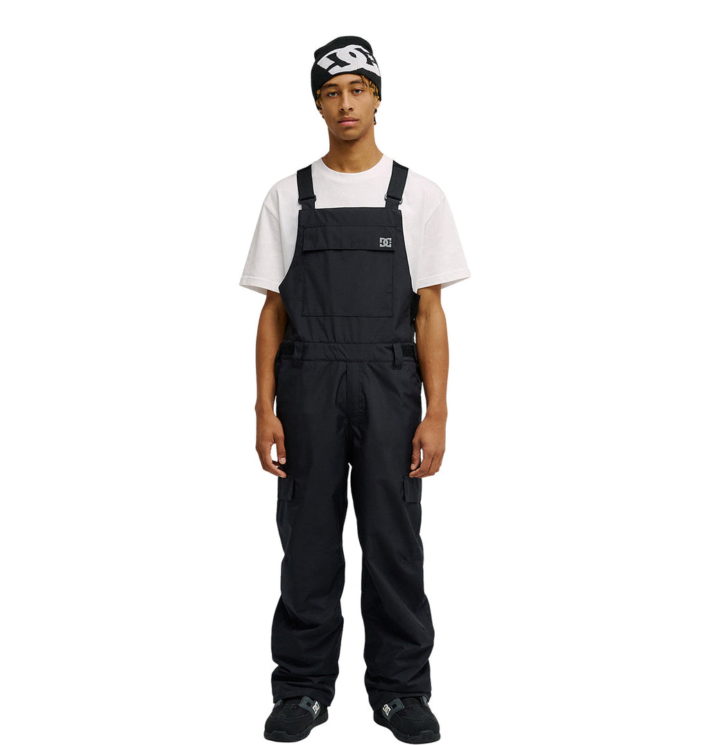 A person stands facing forward against a plain white background, wearing the DC Shoes Docile Bib 10K black snow bib overalls over a white T-shirt, paired with black shoes and a black beanie featuring a white logo.
