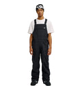 A person stands facing forward against a plain white background, wearing the DC Shoes Docile Bib 10K black snow bib overalls over a white T-shirt, paired with black shoes and a black beanie featuring a white logo.