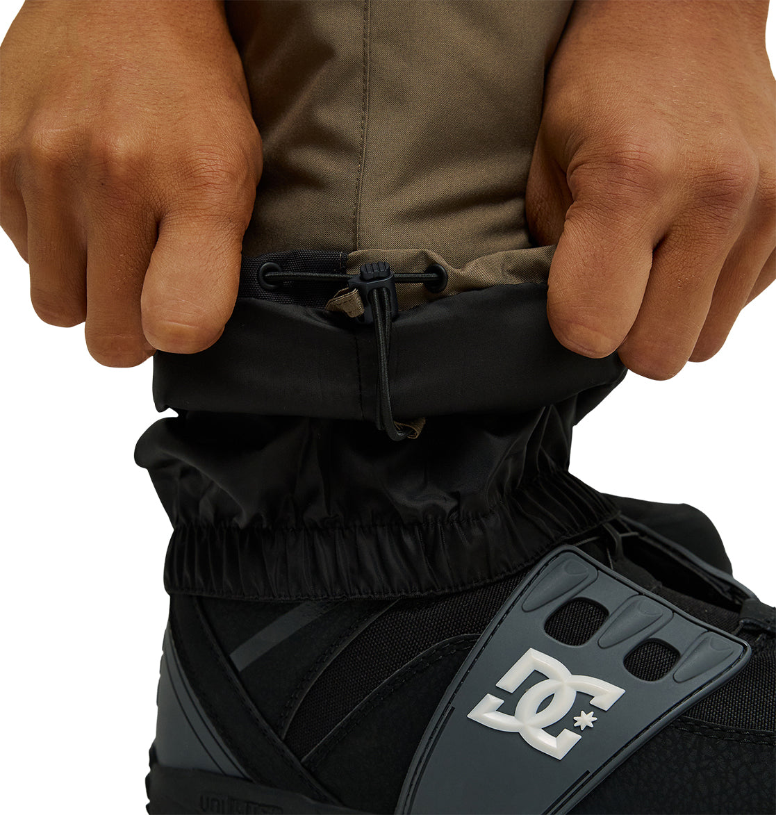 Close-up of hands adjusting the black elastic cuff of the DC Shoes Docile Bib 10K brown snow bib pants, offering breathable protection and waterproof insulation, over a black and gray DC snowboard boot.