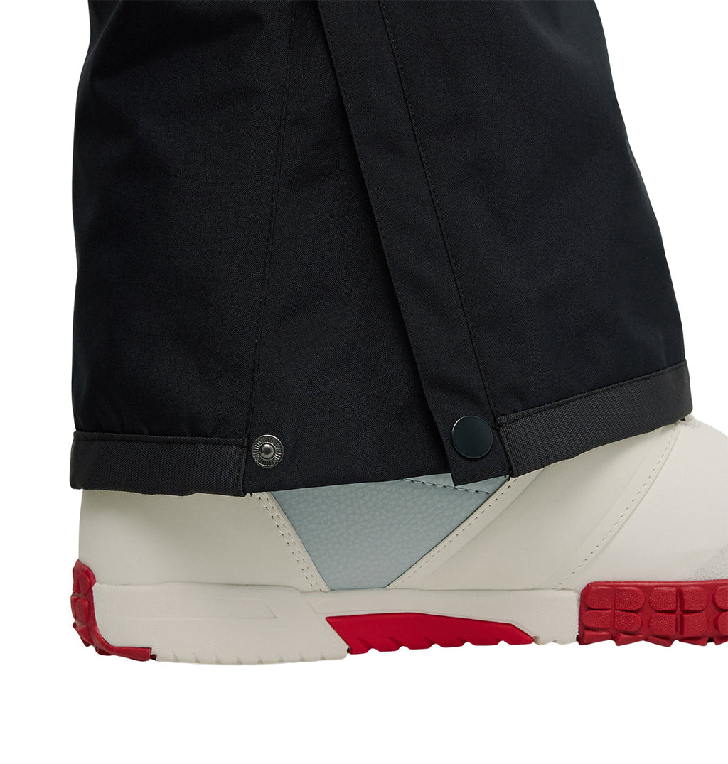 Close-up of a person wearing DC Shoes Banshee Pants, black insulated pants designed for optimal snow performance. The pants feature snap buttons near the ankle and are worn over white and gray winter boots with red soles.