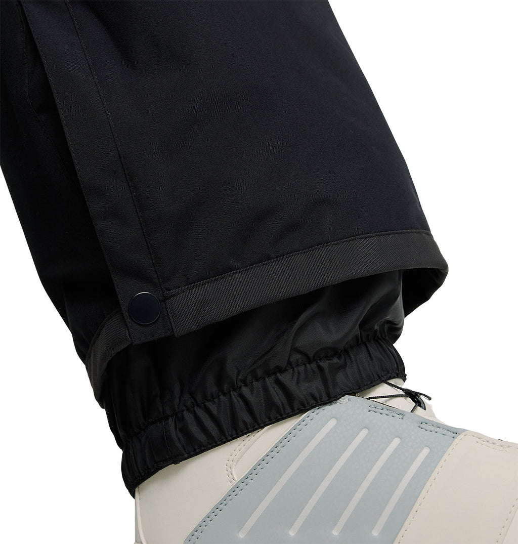 Close-up of a person wearing DC Shoes Banshee Pants, featuring black insulated snow pants with an elastic cuff and a button near the ankle for extra protection over white and gray winter boots. The Banshee Pants offer added warmth and waterproof performance in snowy conditions.