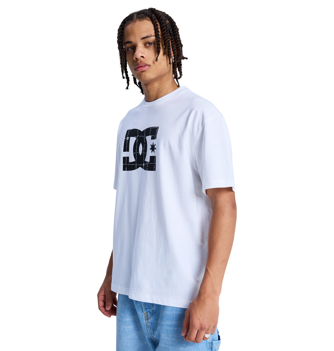 A young man with braided hair wears the Reebok DC Star Filled Hss, a loose white T-shirt with a bold black DC logo on the chest, paired with light blue jeans, standing against a plain white background.