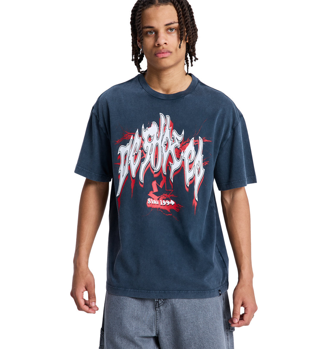 A person with braided hair wears a loose-fitting, dark blue DC Death Machine Hss graphic T-shirt by Reebok with white and red lettering and designs, paired with light blue shorts, standing against a plain white background.