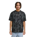 A young person with short, curly hair wears the Reebok DC Liquid Fuego Hss t-shirt—black with a gray flame pattern and "Reebok" printed in white on the chest—paired with light blue jeans, standing against a plain white background.