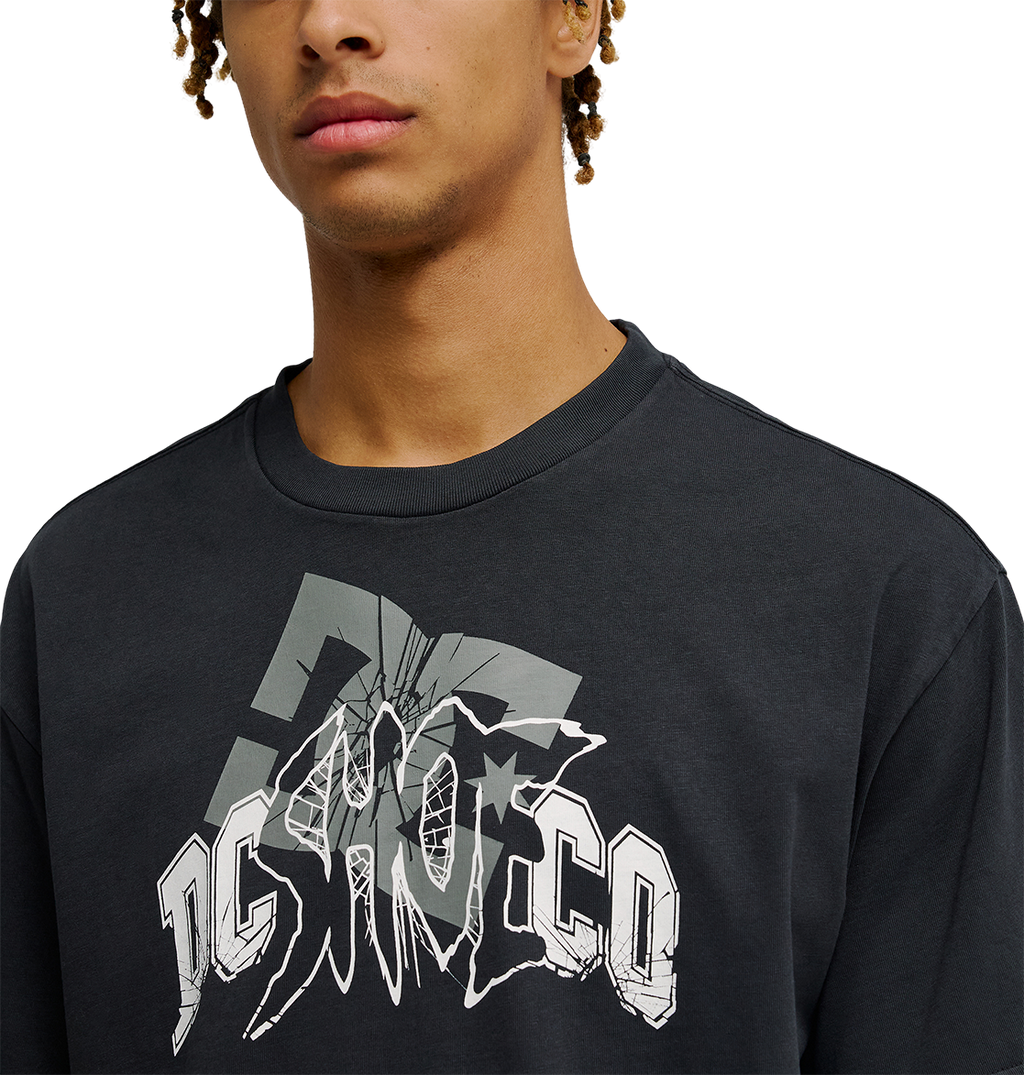 A person wearing the Men's DC Speratic Tee by DC Shoes—a black, relaxed-fit skate t-shirt featuring a bold, stylized gray and white "DC SHOCO" cracked-effect graphic. The heritage-inspired artwork stands out while only the lower half of the face is visible.