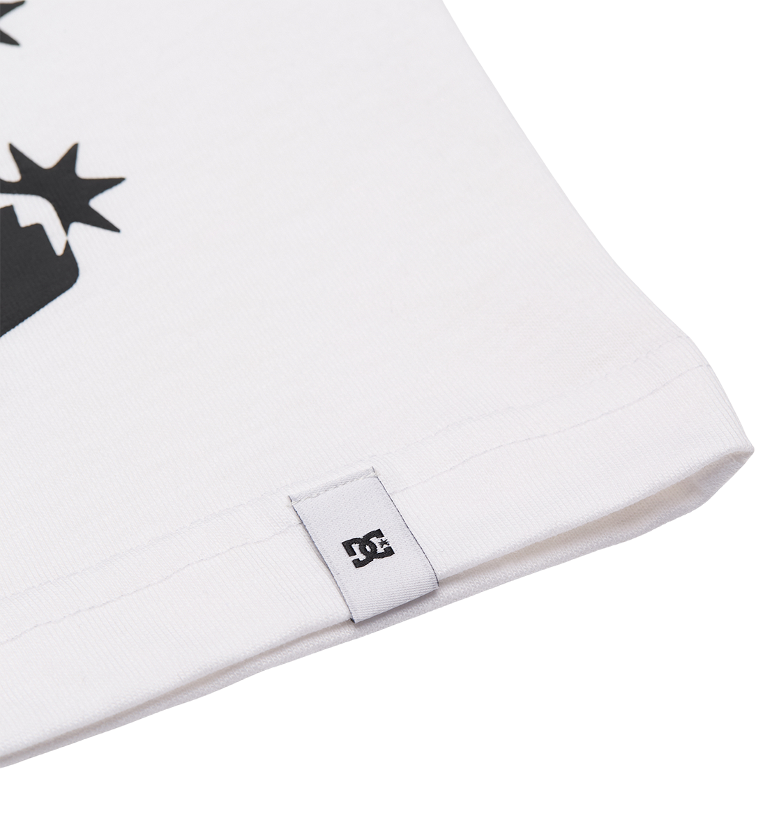Close-up of the hem on the Men's DC Side Flame Tee by DC Shoes, featuring a small white tag with the black DC Shoes logo. The relaxed fit shirt shows part of its black graphic design against a plain white background.
