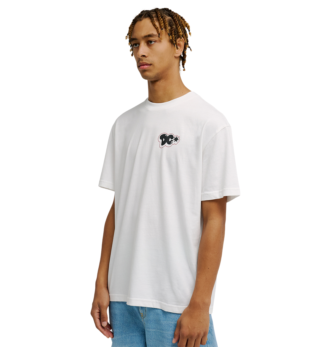 A person with short, curly hair wears the Men's DC Side Flame Tee by DC Shoes—a relaxed fit white t-shirt featuring a small black graphic skate tee design on the chest—paired with light blue jeans, standing against a plain white background.