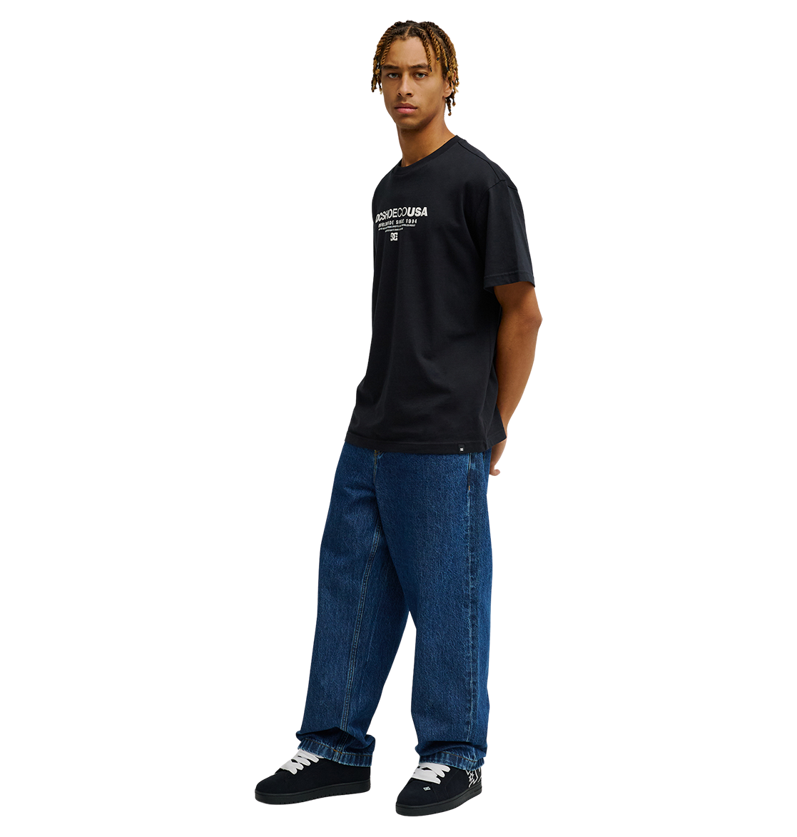 A person stands against a white background wearing the Men's DC Heritage Font Tee by DC Shoes, styled in black with a bold heritage font graphic. They pair it with relaxed-fit loose blue jeans and black sneakers. With short braided hair, they look slightly to the side, hands tucked in their pockets.