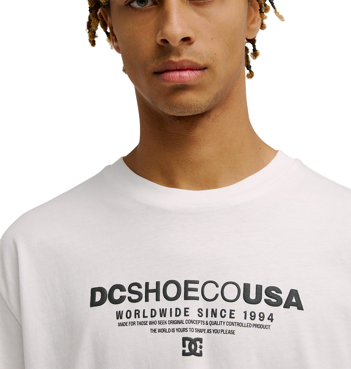 A person wearing the Men's DC Heritage Font Tee by DC Shoes, a white graphic skate t-shirt featuring bold black text that reads "WORLDWIDE SINCE 1994" with additional smaller lettering beneath. The photo shows only the lower face and upper torso, drawing attention to the tee's relaxed fit and classic skate style.