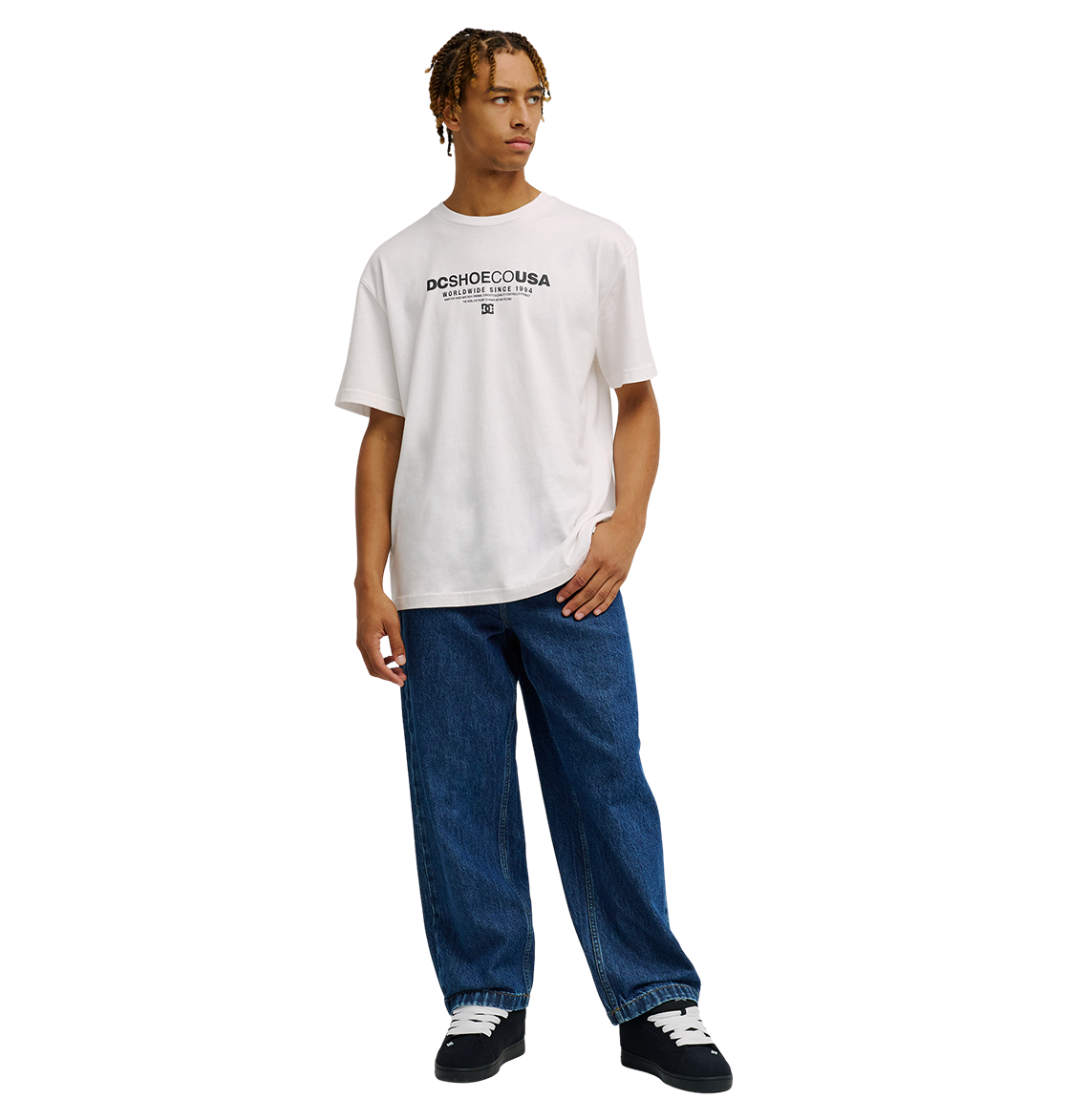 A person stands against a plain white background, wearing the Men's DC Heritage Font Tee by DC Shoes in a relaxed fit with a white graphic design, paired with loose blue jeans and black sneakers with white soles.