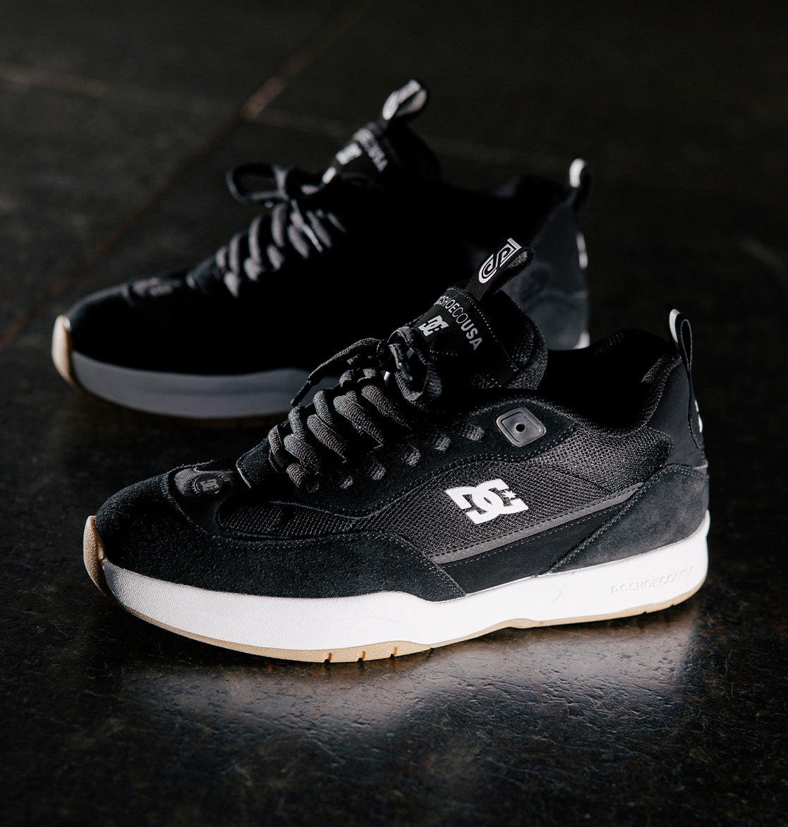 A pair of Men's JS-2 Shanahan Pro Skate Shoes by DC Shoes in black with white soles and branding, inspired by DC heritage, are displayed on a dark surface with one shoe slightly ahead of the other.